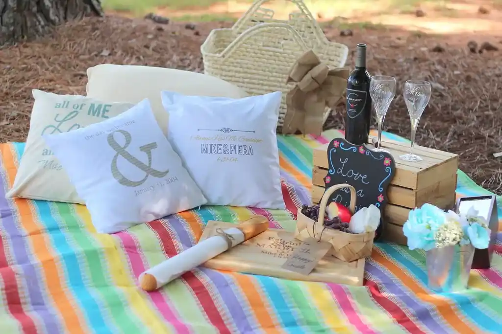 A striped picnic blanket laid out under pine trees, with cushions, a basket, a bottle and two glasses set on it.