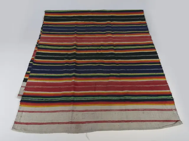 A flat-woven rug in narrow multicoloured stripes laid out flat on a white background.