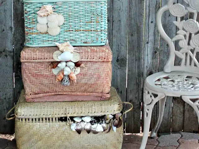 Three lidded wicker hampers, turquoise, pink and natural, each trimmed with sea shells, stacked beside a white iron garden chair.